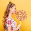 Beautiful young woman addicted to sugar with playful look eating huge candy and smiling. Stylish girl with blonde curly hair. Portrait of attractive lady with big lollypop, yellow wall on background.