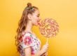 Beautiful young woman addicted to sugar with playful look eating huge candy and smiling. Stylish girl with blonde curly hair. Portrait of attractive lady with big lollypop, yellow wall on background.