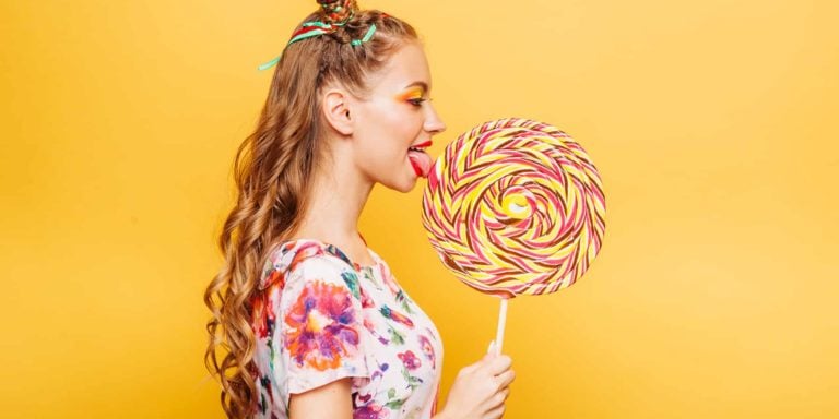 Beautiful young woman addicted to sugar with playful look eating huge candy and smiling. Stylish girl with blonde curly hair. Portrait of attractive lady with big lollypop, yellow wall on background.