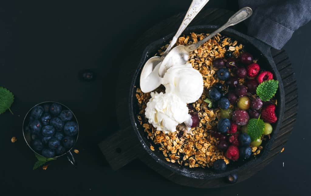 Oat granola crumble with fresh berries, seeds and ice-cream in iron skillet pan on dark wooden board over black backdrop