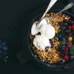 Oat granola crumble with fresh berries, seeds and ice-cream in iron skillet pan on dark wooden board over black backdrop