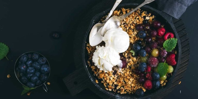 Oat granola crumble with fresh berries, seeds and ice-cream in iron skillet pan on dark wooden board over black backdrop