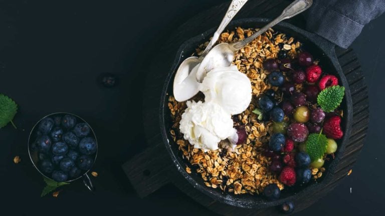 Oat granola crumble with fresh berries, seeds and ice-cream in iron skillet pan on dark wooden board over black backdrop