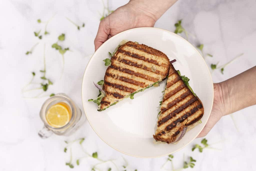two white hands holding a white plate over a white table with a grilled sandwich sliced in half and a lemon slice in a glass on the table