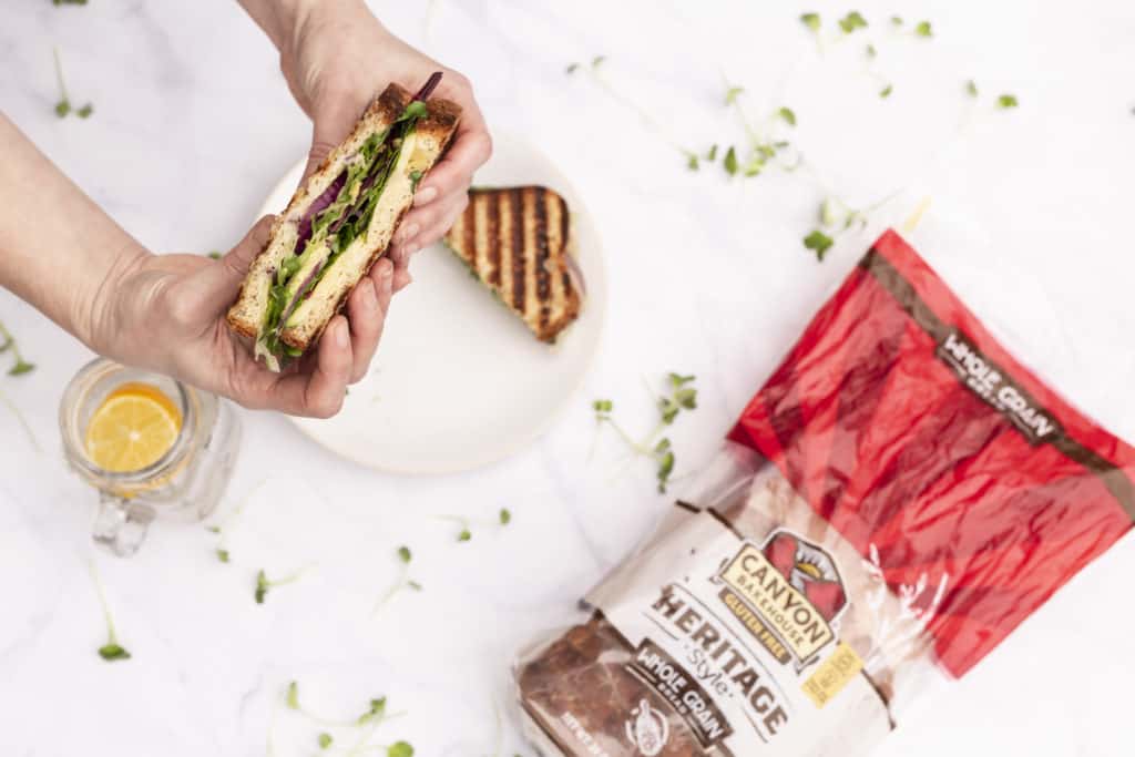 white hands holding one slice of a vegetarian grilled sandwich while other slice is on a white plate with a white background and bread bag to the side