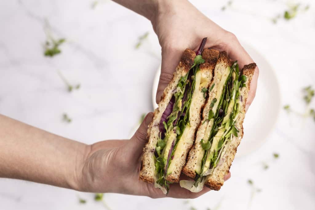 stacked vegetarian sandwich of greens, avocado, zucchini and cheese held together by two white hands