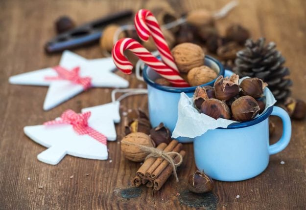 Traditional Christmas foods and decoration. Roasted chestnuts in blue enamel mug, walnuts, cinnamon sticks, candy canes, pine cone on rustic wooden background