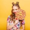 Beautiful young woman with huge candy instead of a head. Big colorful caramel lollypop in hands of woman. Portrait of attractive lady with big lollypop, yellow wall on background.