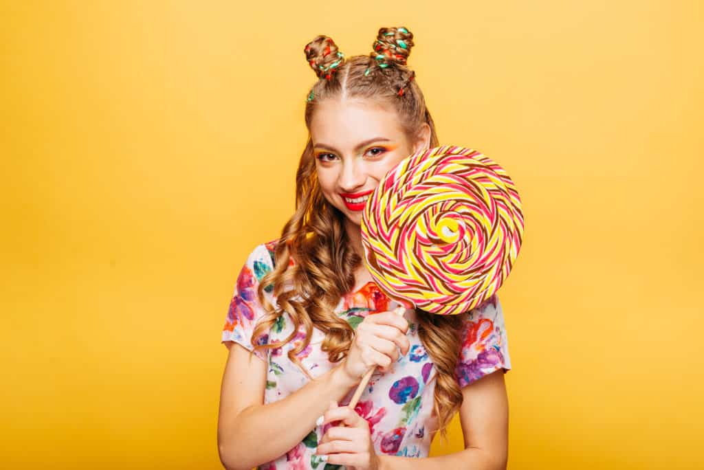 Beautiful young woman with huge candy instead of a head. Big colorful caramel lollypop in hands of woman. Portrait of attractive lady with big lollypop, yellow wall on background.