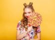 Beautiful young woman with huge candy instead of a head. Big colorful caramel lollypop in hands of woman. Portrait of attractive lady with big lollypop, yellow wall on background.