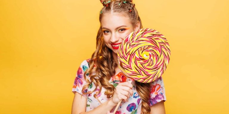 Beautiful young woman with huge candy instead of a head. Big colorful caramel lollypop in hands of woman. Portrait of attractive lady with big lollypop, yellow wall on background.