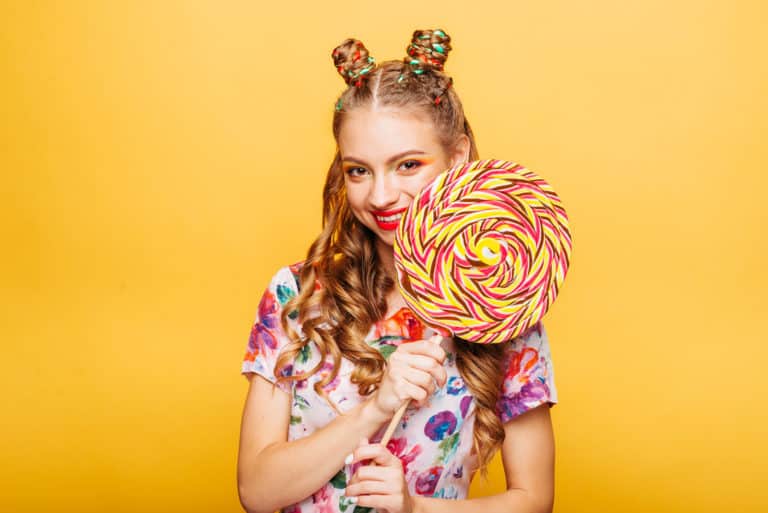 Beautiful young woman with huge candy instead of a head. Big colorful caramel lollypop in hands of woman. Portrait of attractive lady with big lollypop, yellow wall on background.