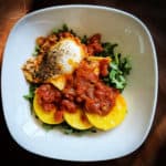 One white plate viewed from above containing one cooked egg, three slices of polenta, red marinara sauce, and some pops of green arugula.