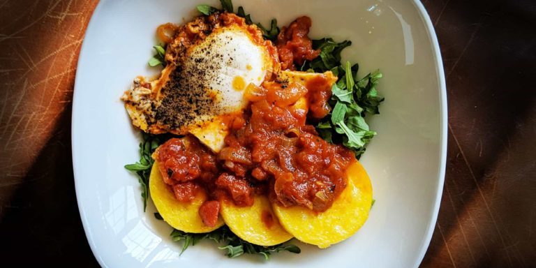 One white plate viewed from above containing one cooked egg, three slices of polenta, red marinara sauce, and some pops of green arugula.