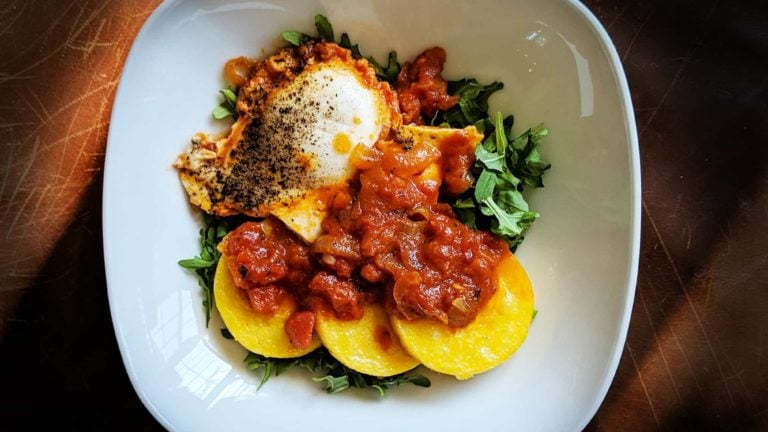 One white plate viewed from above containing one cooked egg, three slices of polenta, red marinara sauce, and some pops of green arugula.