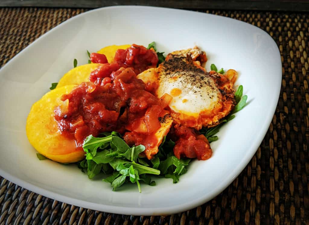 One white plate viewed from above containing one cooked egg, three slices of polenta, red marinara sauce, and some pops of green arugula.