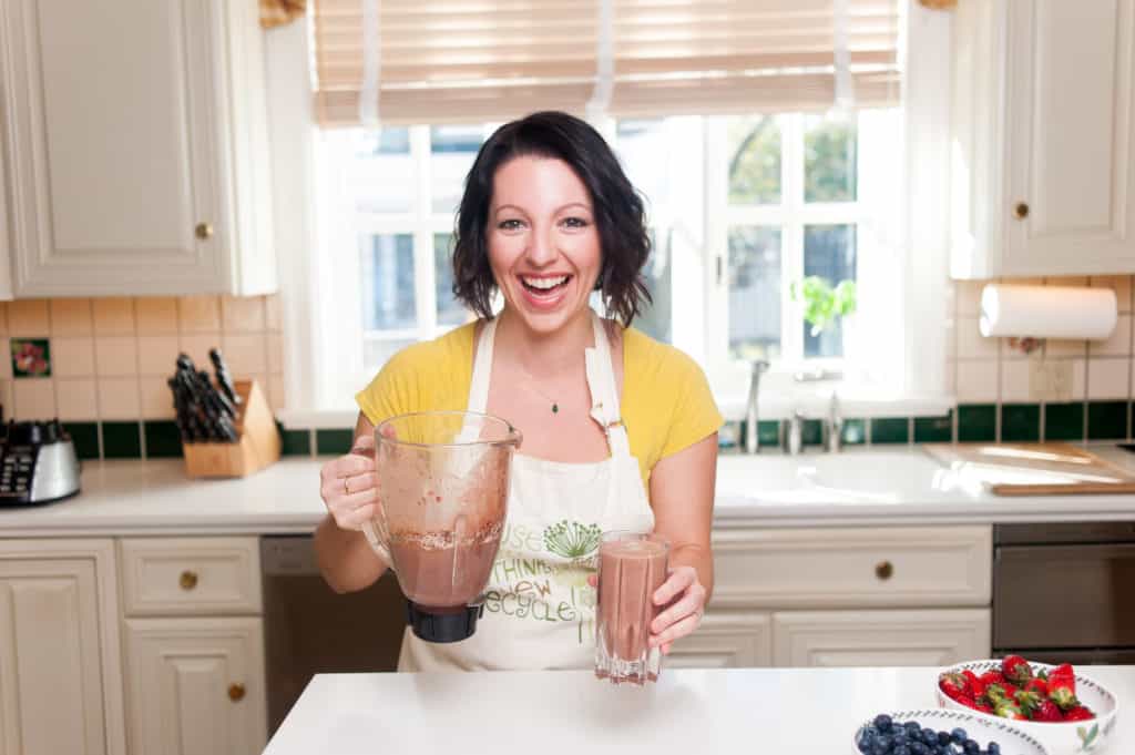 woman laughing holding a blender in one hand and a smoothie in the other hand in a white kitchen