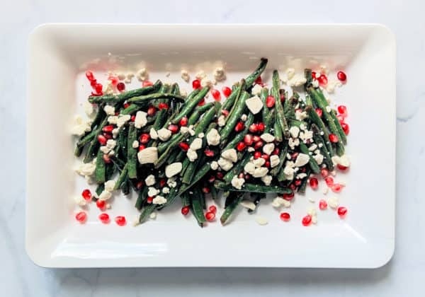 roasted green beans with crumbled goat cheese and pomegranate seeds on a white rectangular plate and light grey background