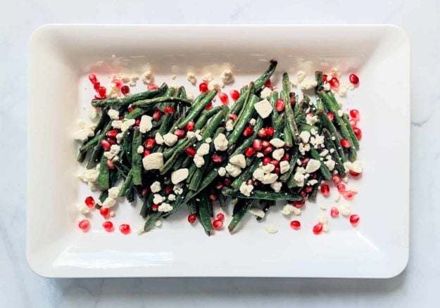 roasted green beans with crumbled goat cheese and pomegranate seeds on a white rectangular plate and light grey background