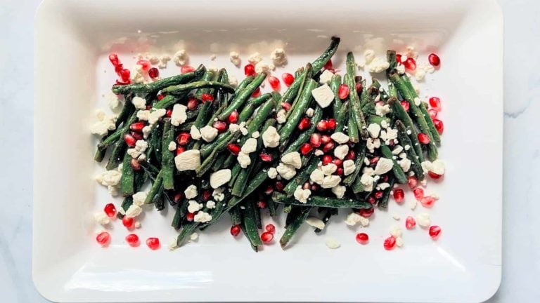 roasted green beans with crumbled goat cheese and pomegranate seeds on a white rectangular plate and light grey background